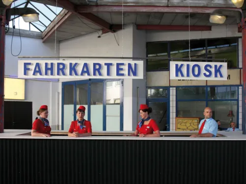 Ein Tresen mit der Beschilderung "Fahrkarten" und "Kiosk". Hinter dem Tresen stehen drei Frauen im Kostüm und ein Mann im kurzärmliges Hemd und mit Krawatte.